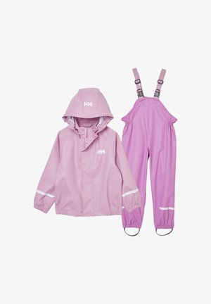 Niña joven sonriente, vestida con chaqueta y pantalones impermeables rosas con tiras reflectantes y botas negras, sobre fondo blanco.