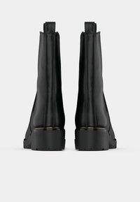 Crne kožne čizme do gležnja s elegantnim dizajnom, koje imaju čvrst potplat i zlatnu akcentnu traku na peti. Glatka tekstura, bez vidljive metalne opreme.
