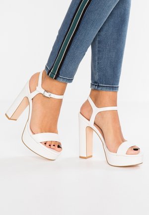 Białe sandały na wysokim obcasie z platformą, z szerokim paskiem na palcach i paskiem na kostkę zapinanym na srebrną klamrę.