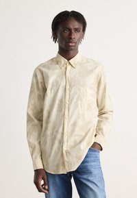 Beige kauluspaita, jossa on texturoitu lehtikuosi, yksi rintatasku ja klassinen kaulus, yhdistettynä sinisiin farkkuihin.
