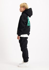 Zwarte hoodie met een grote groene afbeelding op de achterkant, gedragen met bijpassende zwarte joggers en witte sneakers. Gladde stoftextuur, comfortabele pasvorm.