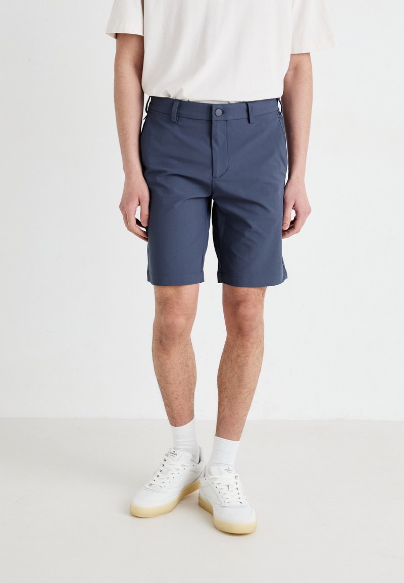 Men's dark blue shorts, tailored fit, made of lightweight fabric, featuring a front button closure and side pockets. Worn with white sneakers.