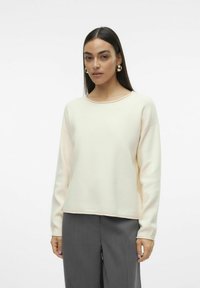 Sweatshirt crème avec un col rond, des épaules tombantes et des manches longues, associé à un pantalon gris. Texture lisse, coupe décontractée.