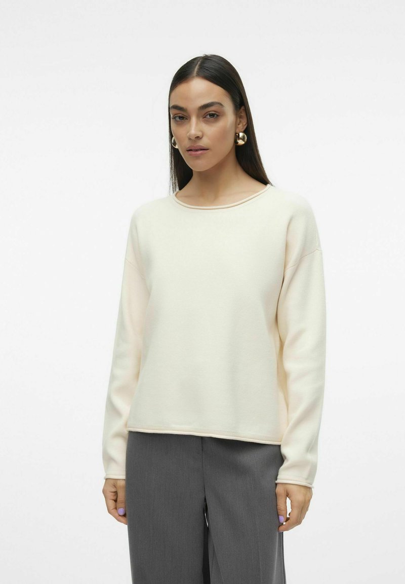 Sweatshirt crème avec un col rond, des épaules tombantes et des manches longues, associé à un pantalon gris. Texture lisse, coupe décontractée.