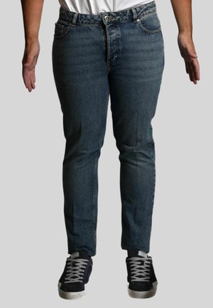 Persona che indossa jeans di denim blu e scarpe da ginnastica nere con lacci bianchi, in piedi con le braccia rilassate ai lati contro uno sfondo semplice.