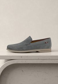 Γκρι δερμάτινο loafer slip-on με ραμμένα διακοσμητικά, μπεζ σόλα και μικρή κόκκινη-λευκή-γκρι υφασμάτινη ετικέτα στο πλάι, τοποθετημένο σε μια μπεζ επιφάνεια.
