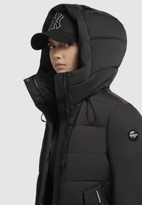 Crna podstavljena jakna s visokim ovratnikom, zatvaranjem na zatvarač i bočnim džepovima. Ima odvojivu kapuljaču i logotip na rukavu.