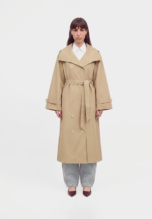 Femme debout portant un trench-coat beige croisé avec ceinture, un jean gris clair et des chaussures pointues foncées sur un fond blanc uni.