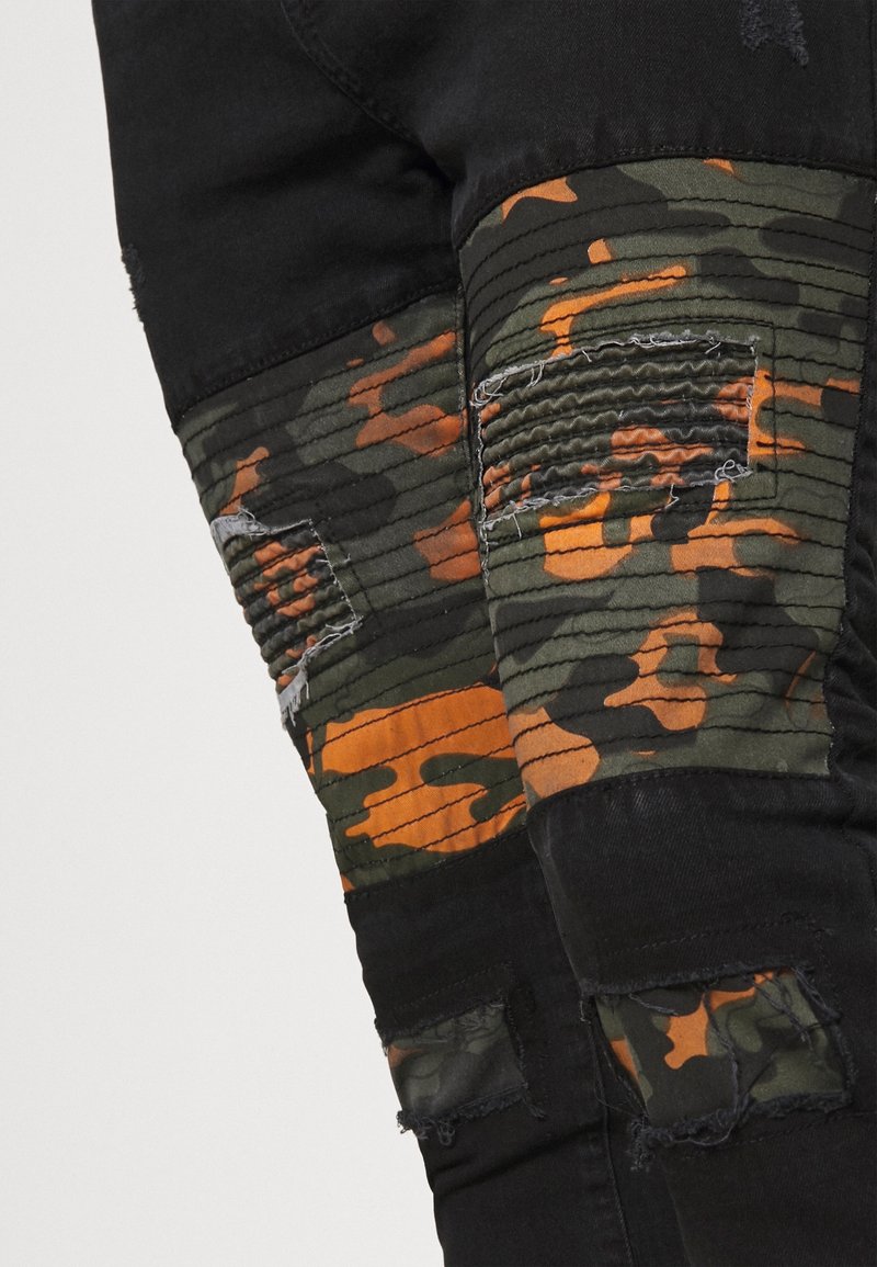 Jeans de mezclilla negra con un panel de camuflaje en verde y naranja, con costuras texturizadas, detalles desgastados en las rodillas y bordes deshilachados.