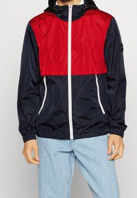Giacca a vento con parte superiore rossa, sezione inferiore blu navy, zip bianche e polsini elastici, realizzata in un materiale leggero e lucido.
