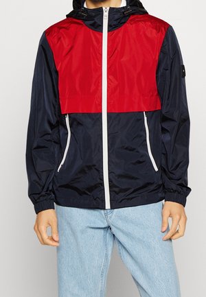 Veste coupe-vent avec une partie supérieure rouge, une section inférieure bleu marine, des zips blancs et des poignets élastiques, fabriquée à partir d'un matériau léger et brillant.