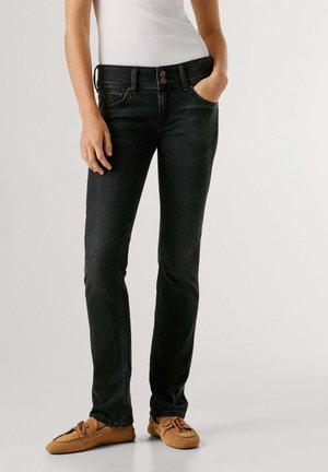 Frau trägt schwarze, eng anliegende Jeans, weißes ärmelloses Oberteil und beige Mokassins aus Wildleder, steht mit einer Hand in der Tasche.