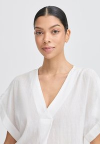 Blouse blanche texturée avec un décolleté en V et des manches courtes. Le tissu présente des rayures subtiles, offrant une apparence légère et aérée.