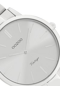 Silver analog watch with a minimalist round face, thin hour markers, and a brushed finish, featuring black brand text and vintage detail.