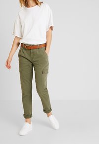 Olive cargo spodnie z bocznymi kieszeniami, podwiniętymi mankietami i brązowym paskiem. Połączone z luźną, białą koszulką i białymi trampkami.