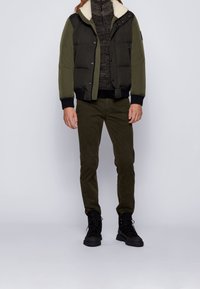 Olivgrön puffjacka med en vit fleecekrage, bärs över en mörk texturerad tröja och olivfärgade byxor. Svarta snörkängor fulländar outfiten.