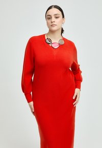 Rotes Strickkleid mit rundem Ausschnitt und langen, leicht puffigen Ärmeln. Kombiniert mit einer mehrfarbigen, klobigen Halskette. Glatte Textur.