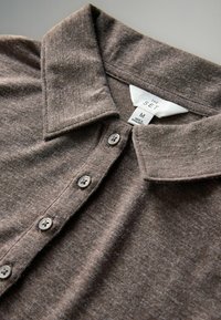 Brūns adīts polo krekls ar mīkstu tekstūru, ar apkakles kaklu, piecām pelēkām pogām un etiķeti, kas norāda izmēru M, ražots Bangladešā.