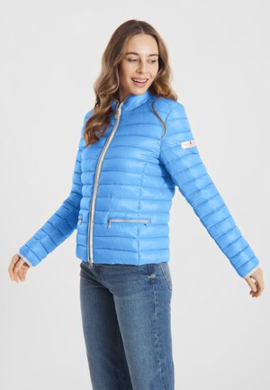 Vrouw die glimlacht, draagt een lichtblauwe gewatteerde jas met ritssluiting en blauwe spijkerbroek, staand tegen een effen witte achtergrond.