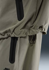 Khaki jacka med åtdragna ärmar, svarta dragkedjefunktioner och mjukt, lätt tyg. Inkluderar snörning för justerbar passform.