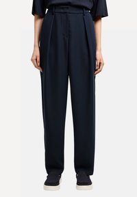 Pantalones de talle alto en color navy con plisados, confeccionados en una tela suave. Presentan bolsillos laterales y un corte ajustado, combinados con zapatos oscuros.