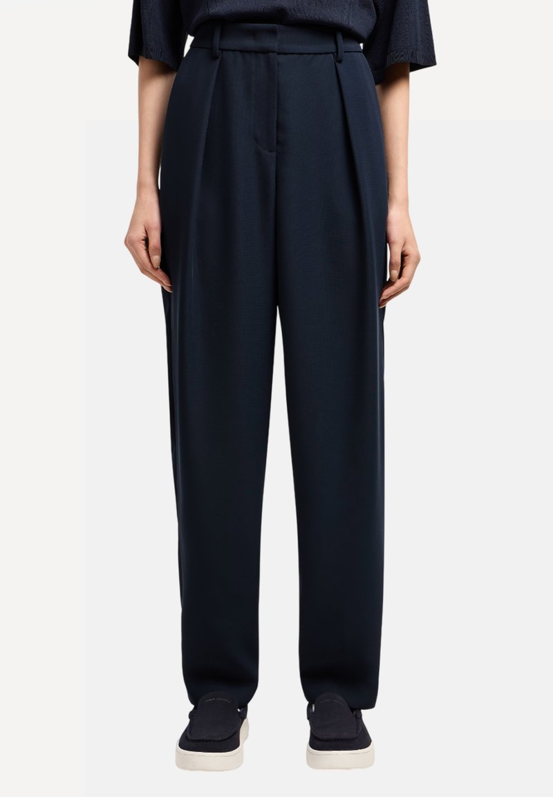 Pantalones de talle alto en color navy con plisados, confeccionados en una tela suave. Presentan bolsillos laterales y un corte ajustado, combinados con zapatos oscuros.