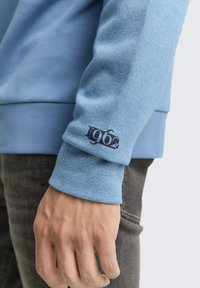 Sweat-shirt bleu clair avec une finition texturée. Comprend un poignet avec "1962" en bleu marine. Le tissu a un léger brillant qui complète le design ajusté.