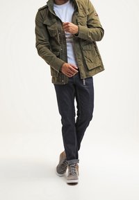 Chaqueta utilitaria verde oliva con múltiples bolsillos, llevada sobre una camiseta blanca, combinada con jeans oscuros y zapatillas gris con acentos marrones.