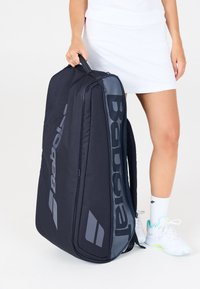 Donna con gonna bianca e scarpe, che tiene in mano una borsa da tennis nera Babolat per il manico su uno sfondo bianco.