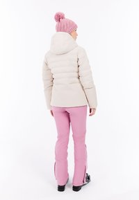 Lichtbeige pufferjack met een capuchon, gecombineerd met roze skibroeken; heeft een geribde roze beanie met een pompon.