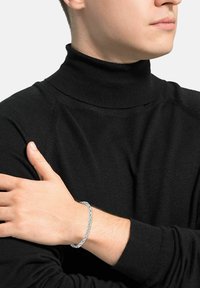 Pulsera de cadena de plata en la muñeca, con eslabones entrelazados. La persona que la lleva puesta viste un suéter negro de cuello alto con una textura de canalé.