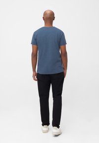 Blauwe katoenen heren T-shirt met korte mouwen, gecombineerd met zwarte jeans en witte sneakers, van achteren bekeken tegen een effen achtergrond.