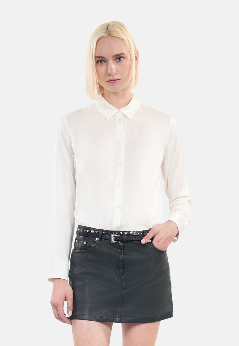 Witte blouse met lange mouwen en een kraag, met een knopenfront; gecombineerd met een zwarte leren minirok met glanzende gespaccenten.