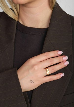 Zlatý prsteň s vlnitým dizajnom na ruke so svetloružovými nechty, opretý o tmavohnedý blejzer s pruhmi. Minimalistická estetika.