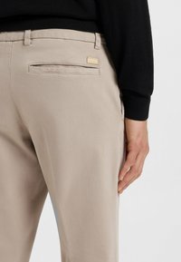 Beżowe spodnie o gładkiej fakturze, z jedną tylną kieszenią i małą złotą metką z logo. Noszone z czarnym swetrem.