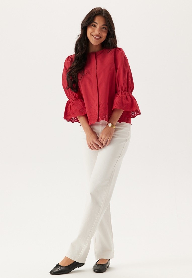 Blusa rossa in cotone con maniche a volants e orlo ricamato, abbinata a pantaloni bianchi e ballerine nere. Stile semplice e casual.