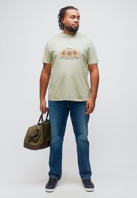 Άνδρας με πράσινη γραφική μπλούζα με ηλιοτρόπια και κρανίο, μπλε τζιν, μαύρα αθλητικά παπούτσια, κρατώντας πράσινη τσάντα duffel, όρθιος, κοιτάζει δεξιά.