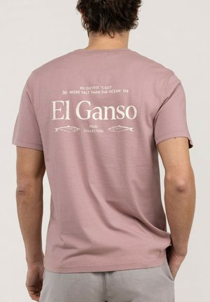 Hombre que lleva una camiseta rosa desteñida con texto blanco y gráficos de peces que dicen "El Ganso SS26 Collection" en la espalda, sobre fondo liso.
