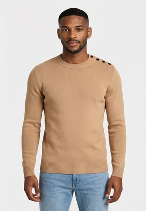 Uomo che indossa un maglione beige a coste con bottoni neri sulla spalla sinistra e jeans azzurri, in piedi davanti a uno sfondo bianco.