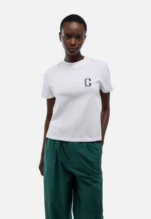 T-shirt blanc en coton avec un col rond et des manches courtes. Présente un petit logo "G" navy sur la poitrine gauche. Associé avec un pantalon vert.