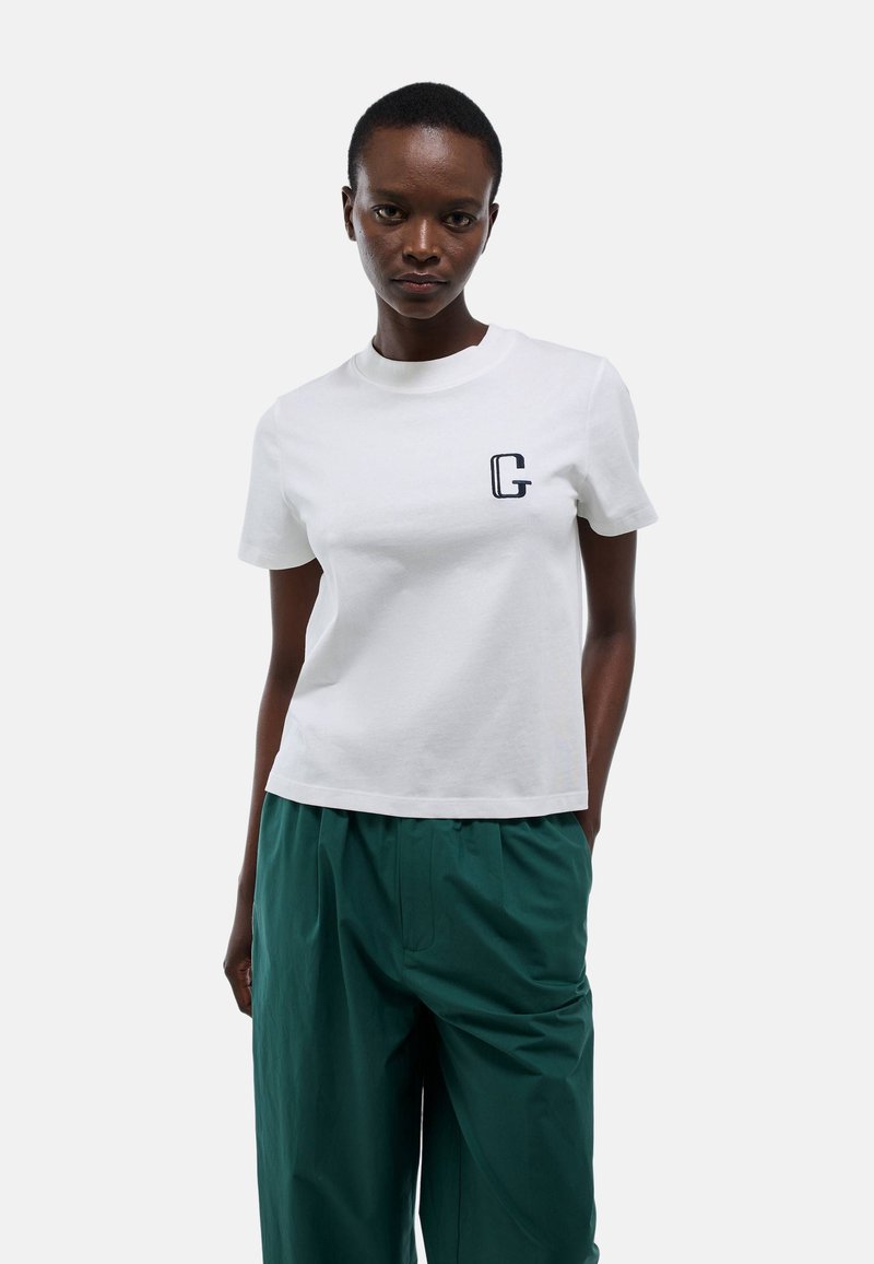 T-shirt blanc en coton avec un col rond et des manches courtes. Présente un petit logo "G" navy sur la poitrine gauche. Associé avec un pantalon vert.