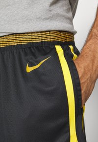 Gros plan d'une personne portant un short de sport noir avec des bandes jaunes, un motif jaune à la taille et un logo Nike jaune, main dans la poche.