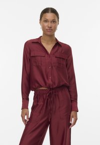 Bordeauxfarbendes Hemd mit Knopfverschluss, zwei Brusttaschen, langen Ärmeln und elastischem Bund. Aus einem glatten, strukturierten Stoff gefertigt. Passende Hose.