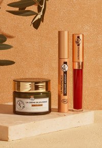 Bocal en verre vert avec couvercle doré étiqueté "LA CRÈME DE JOUVENCE" à côté de deux produits pour les lèvres accentués d'or, un rouge et un transparent.