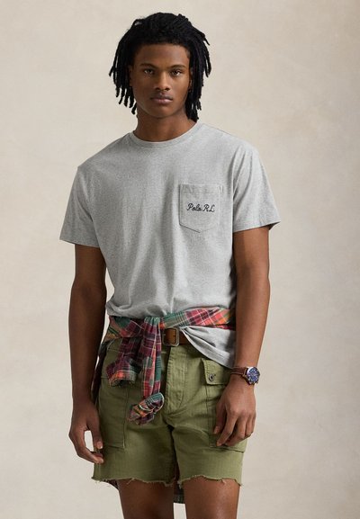 Young man wearing a gray Polo RL T-shirt, green cargo shorts, a plaid shirt tied around waist, and a wristwatch against a plain background.