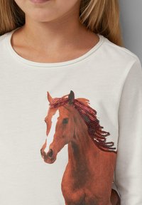 Weißes Langarmhemd mit einem braunen Pferde-Graphic und einem mit roten Pailletten akzentuierten Mähnenteil. Aus weichem Baumwollmaterial mit Rundhalsausschnitt.