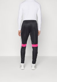 Zwarte sportbroek met roze streepaccenten, witte zijkanten en een trekkoord bij de taille. Het materiaal lijkt glad en glanzend.