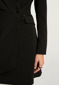 Blazer nero su misura con un design strutturato, cuciture diagonali, chiusura con un bottone singolo e dettaglio della tasca laterale. Tessuto dalla superficie liscia.