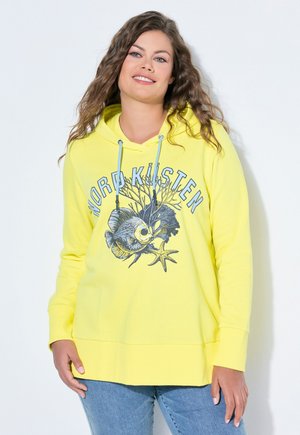 Kvinde med bølget hår iført gul hættetrøje med teksten "Nord Kusten" og grafik af fisk, tang og søstjerne, kombineret med blå jeans.