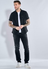 Schwarzes kurzärmliges Hemd mit Knöpfen über einem weißen T-Shirt, kombiniert mit dunklen Jeans und weißen Sneakers. Verfügt über eine dezente Textur und Passform.